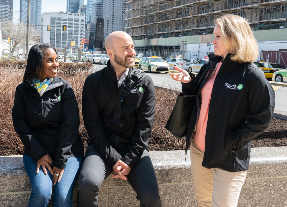 SparkMedical.com® medical affairs team members Group of four Spark Therapeutics Medical Affairs team members outdoors with center two seated on a low wall and outer two standing, looking at one team member and smiling with city skyline in background
